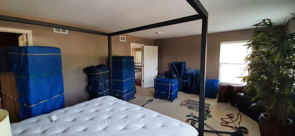 Furniture wrapped in blue moving blankets and strapped, staged in a bedroom around a four-poster bed frame before a move